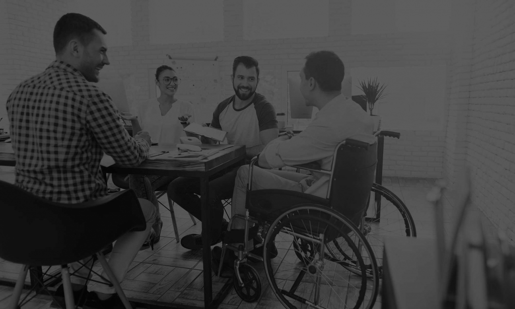 A meeting with a wheelchair user in an office environment. 4 people sit on chairs around a desk, at the top of the table is a wheelchair user.