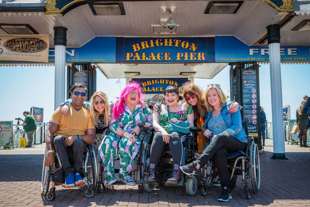 Inclusive Travel. Group of 6 Enhance the UK trainers most of whom are wheelchair users posing together in front of Brighton Pier Sign