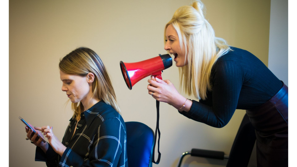 A deaf woman is sitting on a chair looking at her phone. Behind her stands another woman with a red megaphone shouting at the back of her head.