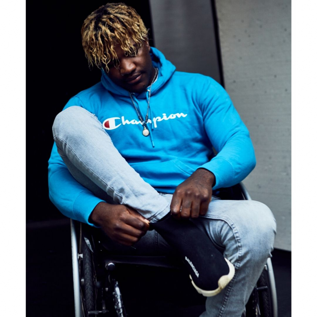 Brinston is pulling up his right sock whilst seated in his wheelchair. He is wearing a bright blue hoodie and has blonde streaks in his dark hair.