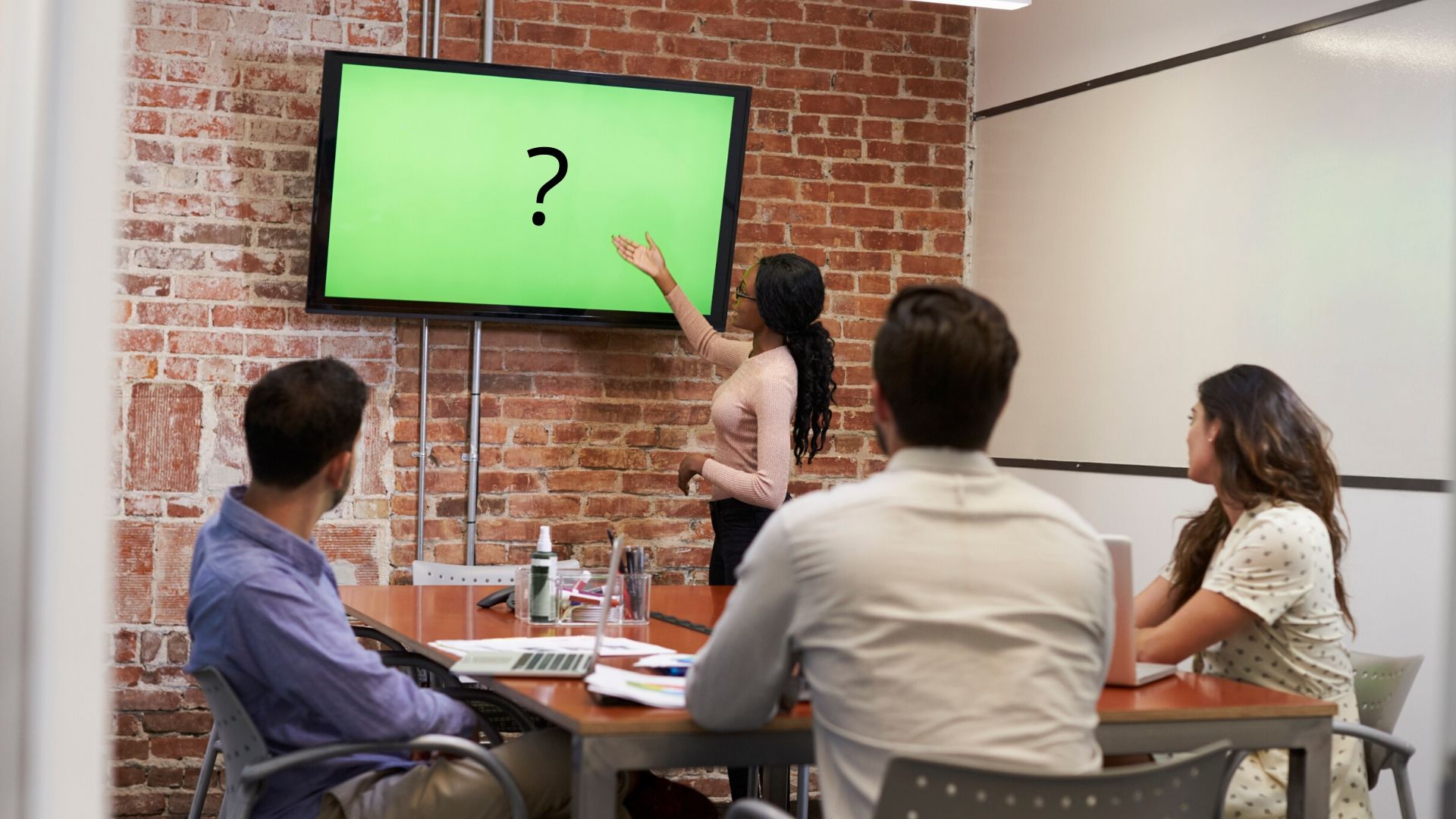 3 people with their backs to the camera are sat at a table watching a person presenting to them. There is a green tv screen with a question mark on it.