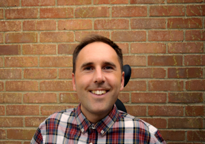 Martyn Sibley is photographed in a tartan print shirt with a big smile on his face. He is in his wheelchair and the background is a brick wall