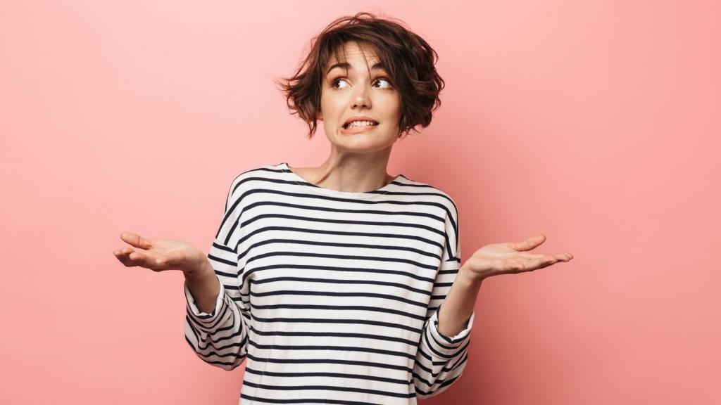 A woman wearing a striped black and white top stands before a pink background and raises her hands in a shrugging motion