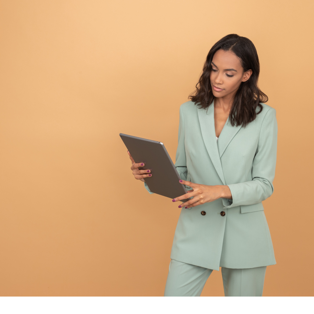 Woman standing in a mint green suit looking at her tablet. She is standing against an orange wall