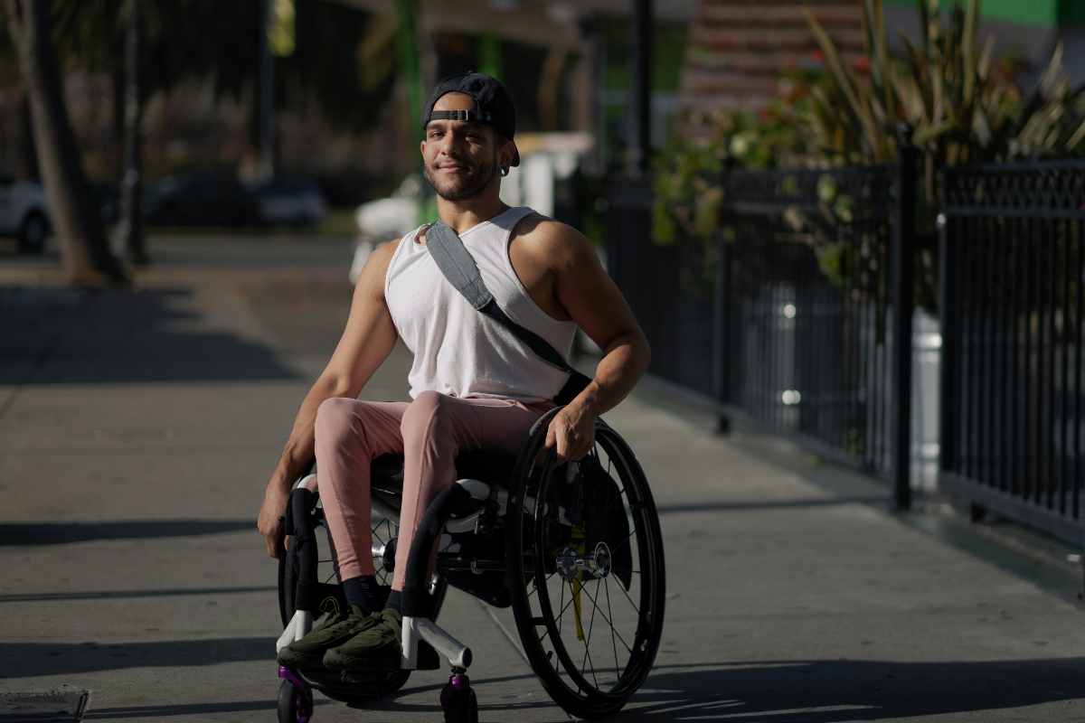 Disability Pride Month: A man is sitting in a wheelchair in the sun. He is wearing a white top, pink pants and a bag on his chest