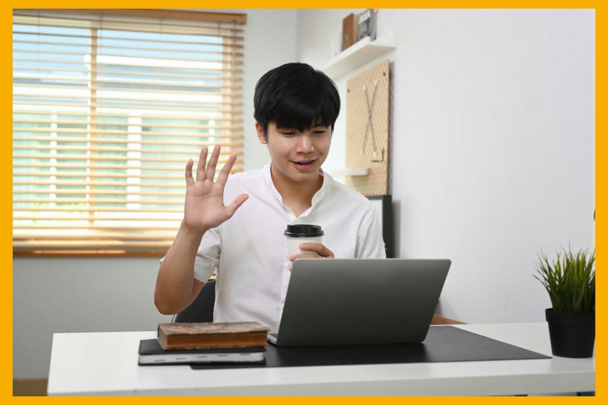 A man is making a sign at a laptop - waving to say hello. He is wearing a white shirt and is sitting at a desk. His laptop is open in front of him